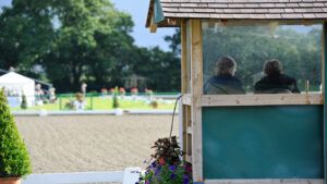 A judges’ box pictured from behind with a judge and their writer sitting inside facing away from the camera, with an outdoor dressage arena behind.