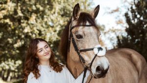 A lady looks lovingly at her dun pony who is wearing a bridle with a crossed noseband