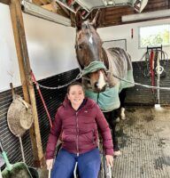 Saffron Cresswell pictured in her wheelchair in front of a horse cross tied in a grooming stall wearing a rug.