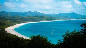 Wide shot of a bay with yachts bobbing about in it. The sand wraps around in a curve and there are mountains behind.