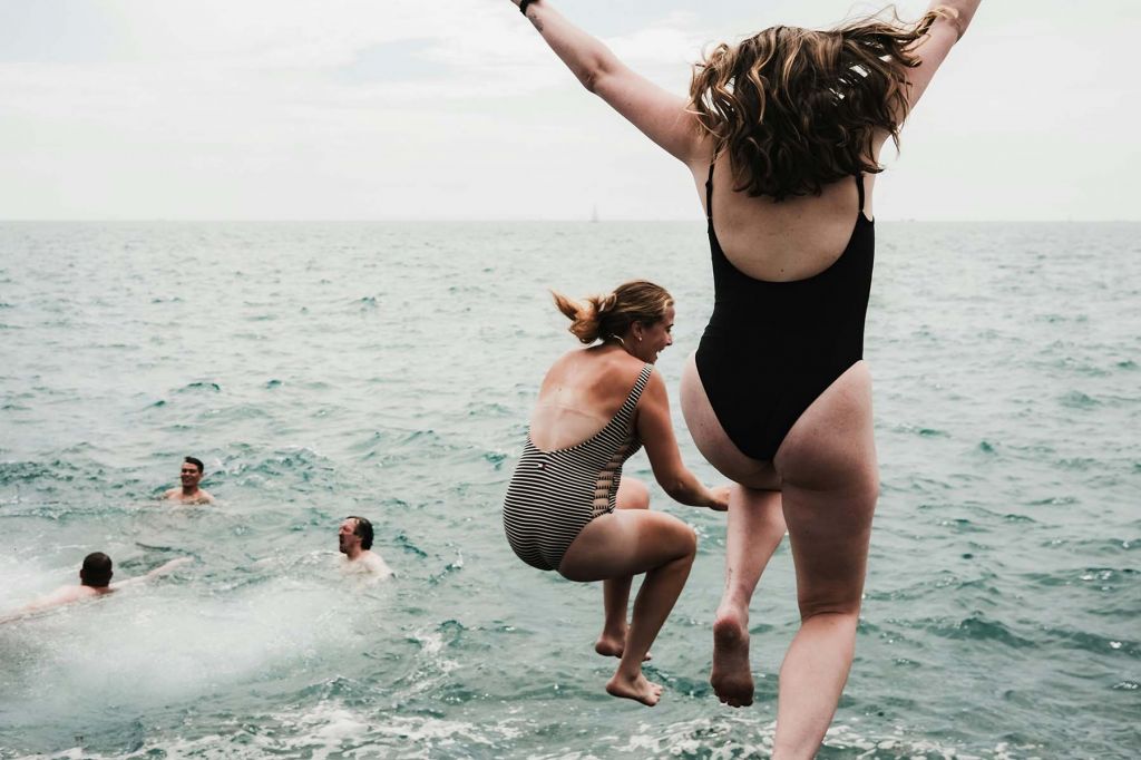 Two middle-age women are seen from behind, jumping into the ocean. Three men are seen already in the water.