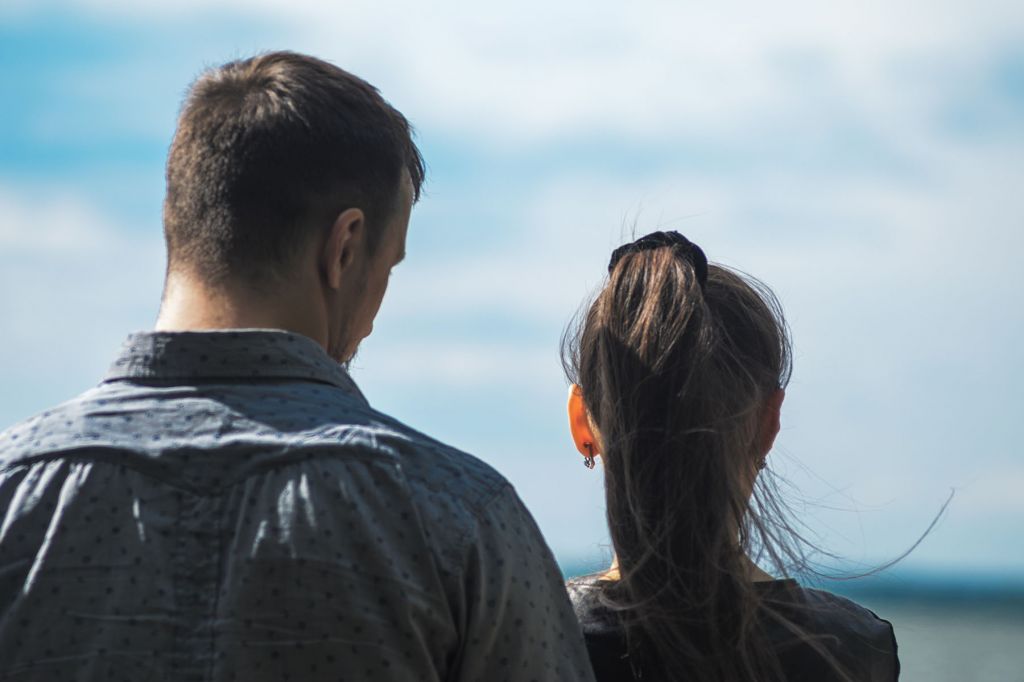 A photograph of a woman and man seen from behind. They're standing in front of a blue sky. The man is looking towards the woman.