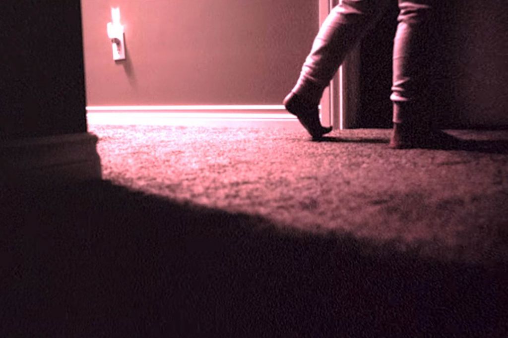 A still from Skinamarink. A close up of a child's legs in pyjamas, walking past shadows and a nightlight in low, red-tinged light.