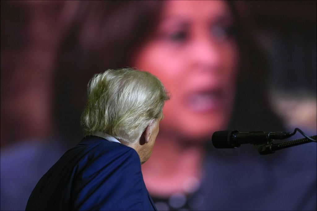 The back of Trump's head as he watches a video of Kamala Harris on a big screen. that is much larger than he is.