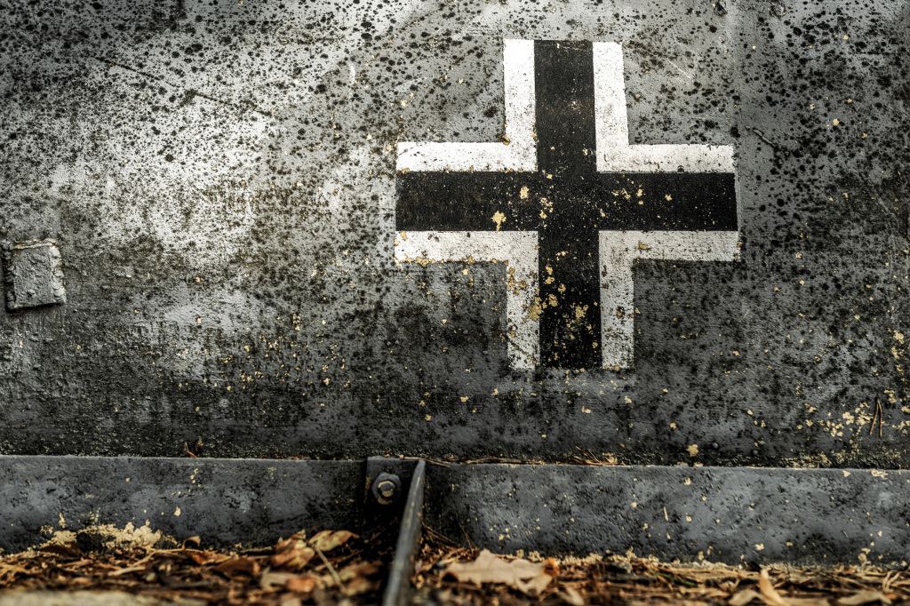 A black-white cross, the emblem of the Wehrmacht, on a grey concrete wall. Below it are dead leaves