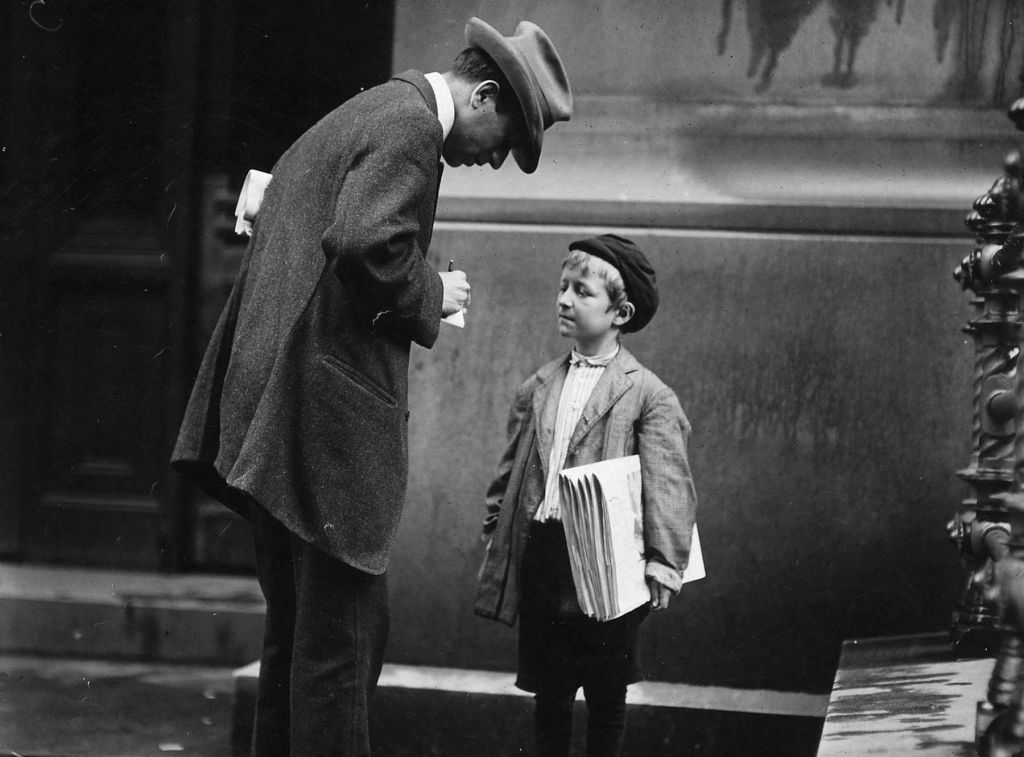 In a black-and-white photo from 1910, a man in an overcoat and hat stoops while writing on a small pad of paper while speaking to a small news boy who holds a stack of papers.