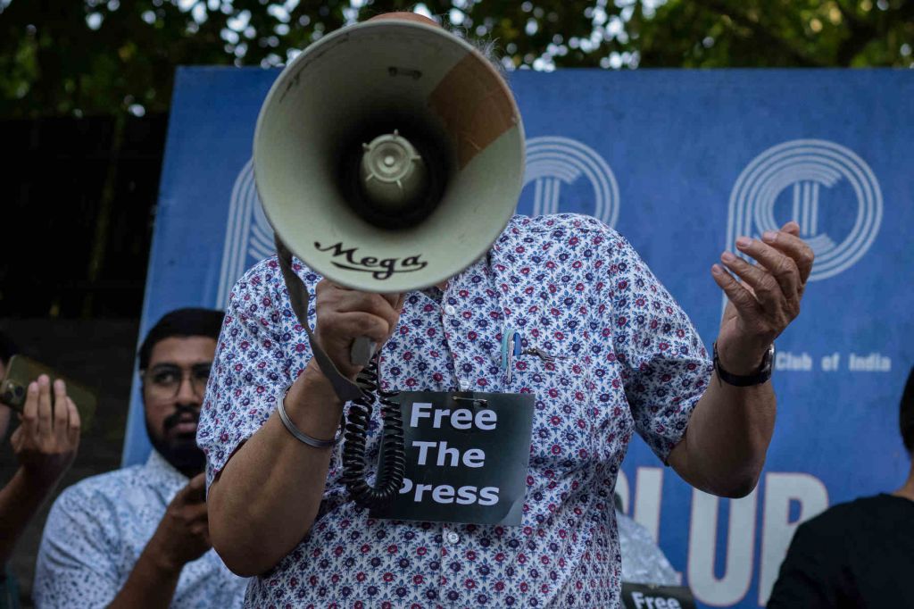 A man wearing a blue shirt and a Free the Press sign pinned to his shirt holds a megaphone that blocks his face.