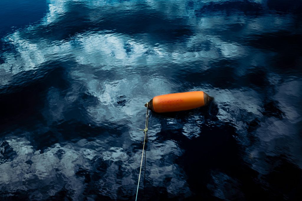 An orange buoy fastened to a long line of rope floats on the surface of dark blue water reflecting clouds