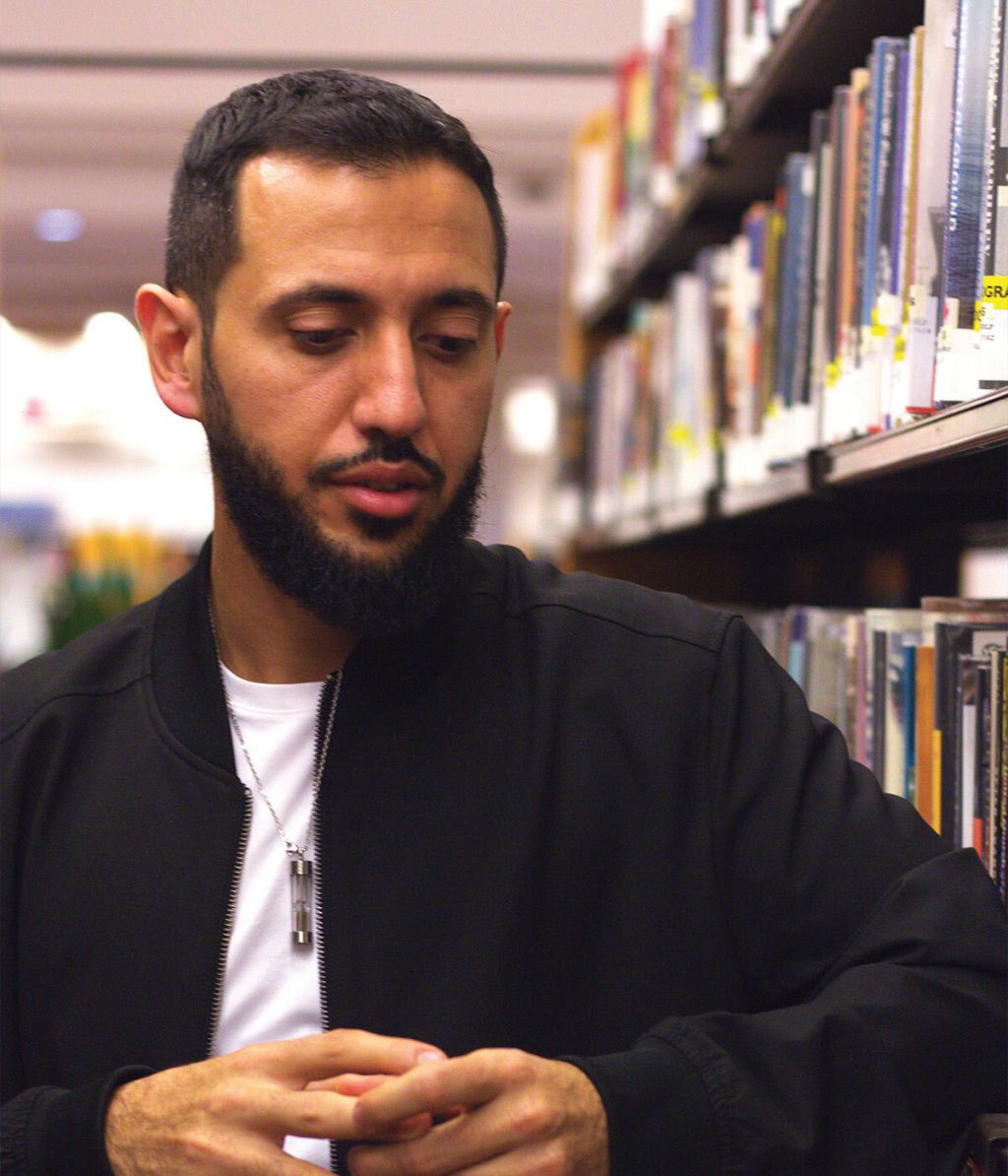 In front of a library bookcase, a man wearing a black jacket over a white T-shirt folds his hands.