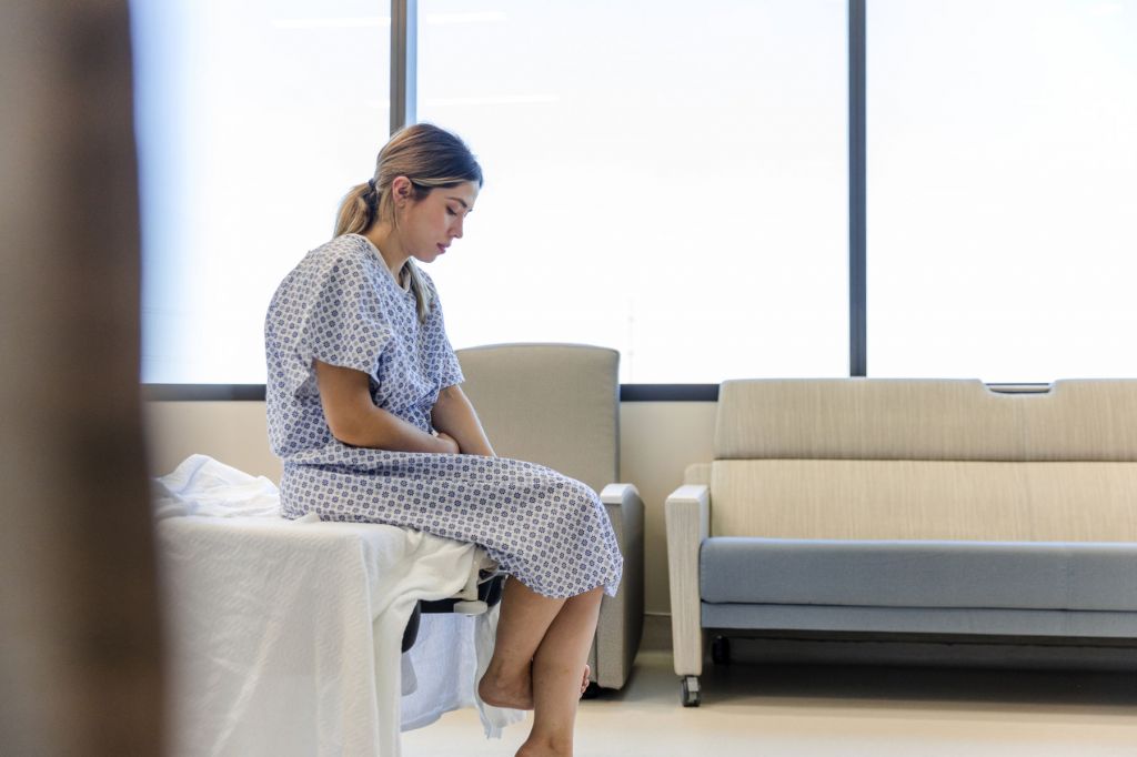 A young woman wearing a hospital gown sits on a bed, looking down at the ground