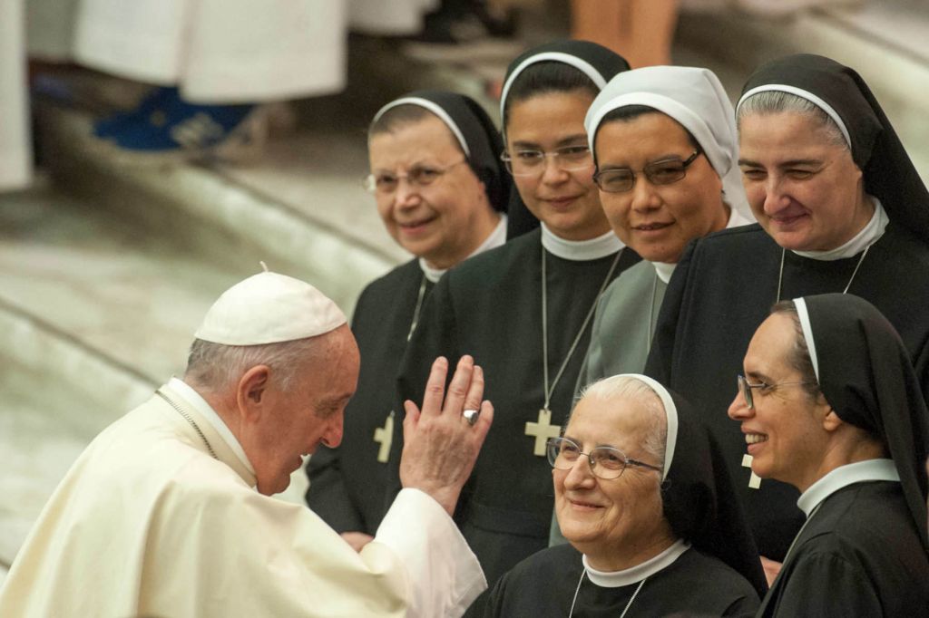 Pope Francis, wearing white, raises his hand to bless a smiling group of nuns, dressed in black, grey, and white habits.