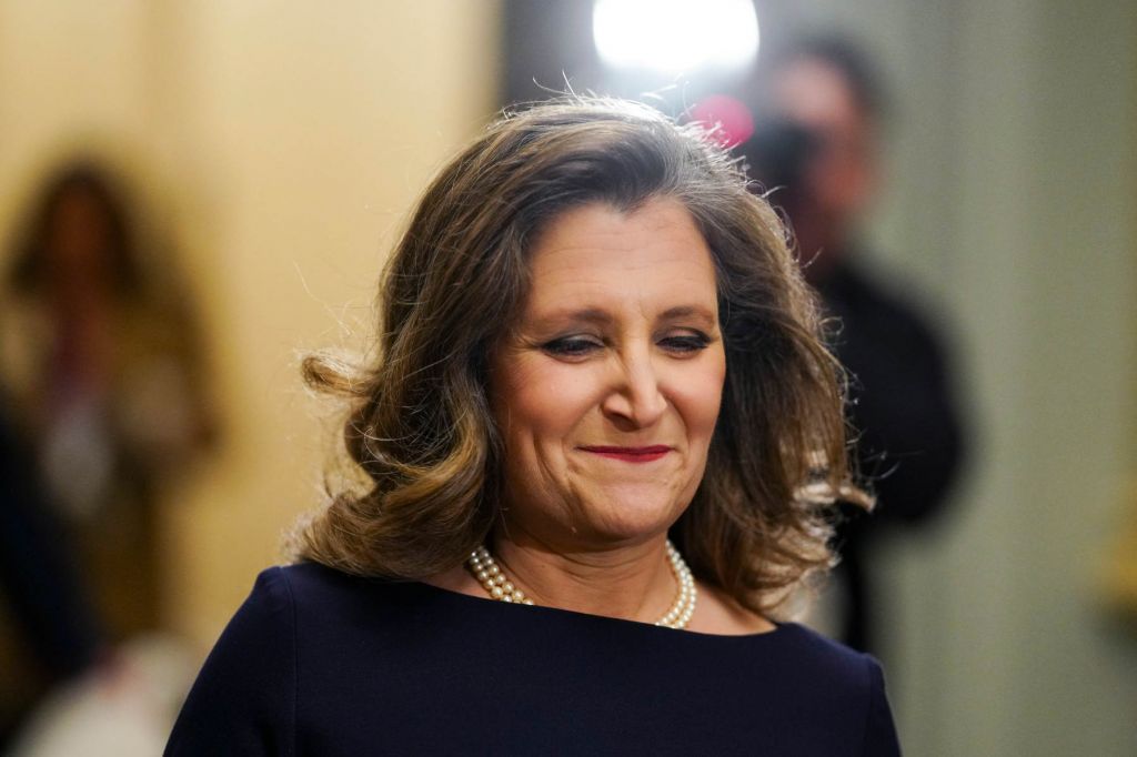 Chrystia Freeland wearing pearls and a black dress, smiles. A camera flash and person holding a camera are in the background