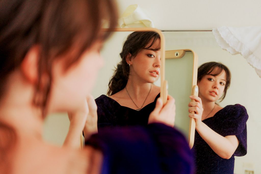 In a photo, a woman in a dark purple dress holds up a mirror to another mirror, so that the smaller mirror and her reflection appear an additional two times.