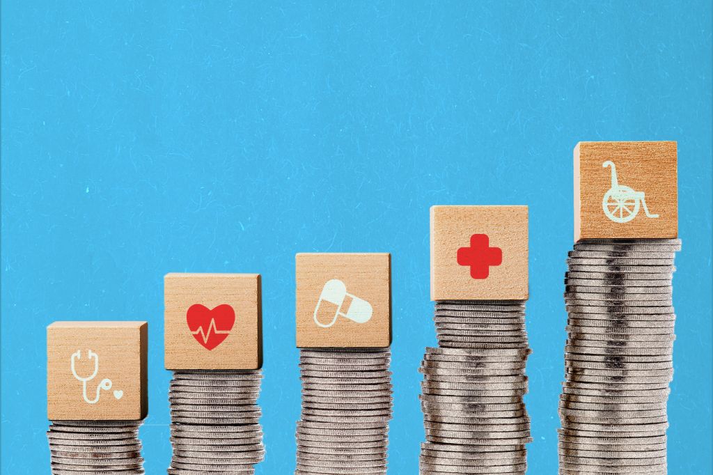 A photo illustration of stacks of coins against a blue background. Atop each stack is a block with a different medical symbol: a stethoscope, heart, pills, red cross, and wheel chair