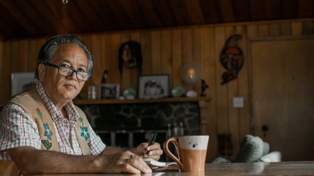 A Cree man with glasses sitting at a wooden table wearing a vest and a button-down. He is holding a pen to a notebook and there's a mug on the table. 