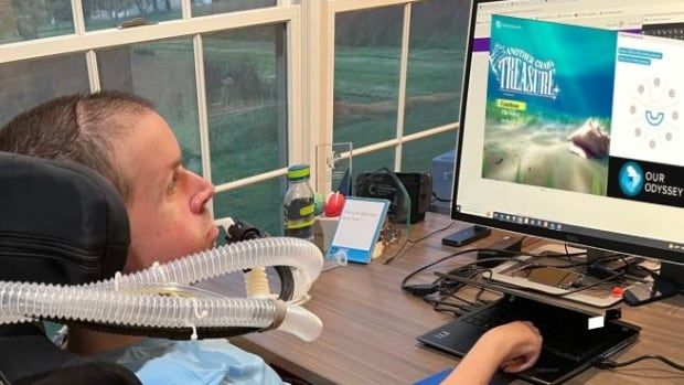 A person in a wheelchair using an assistive breathing device plays a video game on a desktop computer