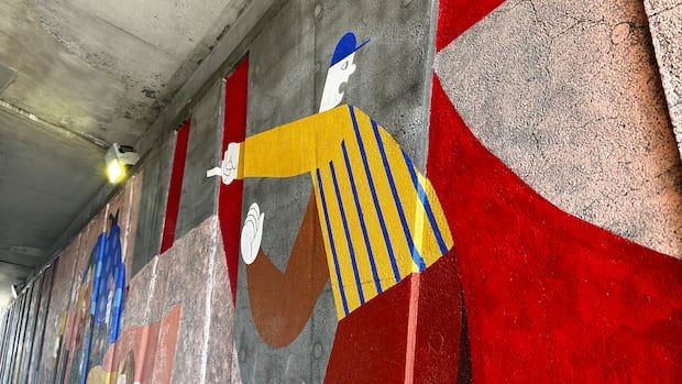 a mural in a concrete underpass depicts a man in a blue hat and yellow shirt pointing, with red motifs behind