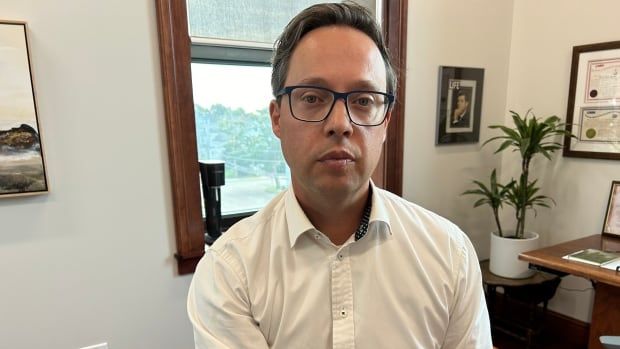 A man with short dark hair and glasses wearing a white dress shirt sitting in an office.