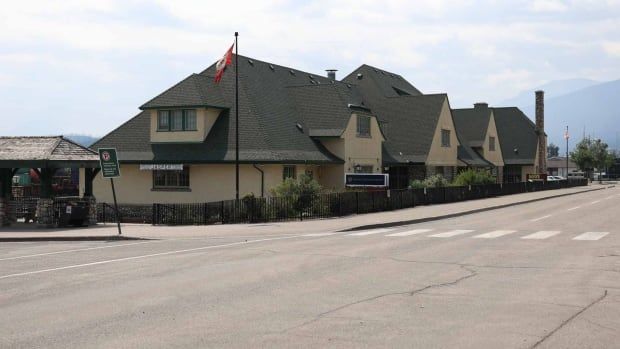 A long beige building with a steeply sloped greenish roof is shown from the street. A flagpole flies the Canadian flag in front.