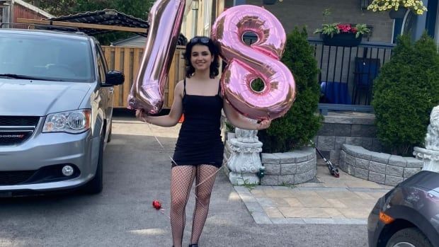 A young woman in a black dress smiles as she holds two pink balloons in the shape of the numbers 1 and 8