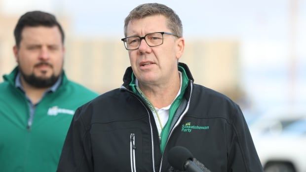 A man stands at a podium. The man is wearing a white shirt with a black jacket over top. The jacket has the phrase "Saskatchewan Party" printed on it.