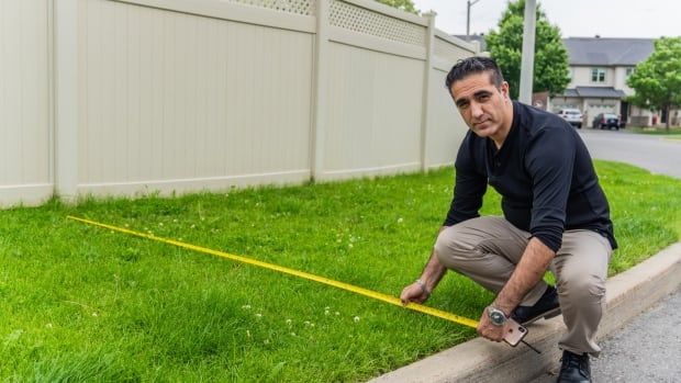 A man measures a white fence to the curb with a measuring tape.