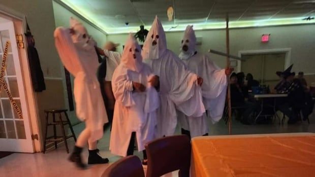 A group of people in white KKK costumes with a cross are seen in a fire hall. 