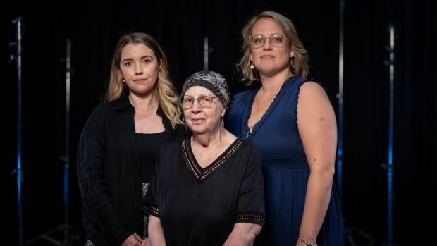 Three women stand side by side, the middle woman sitting.