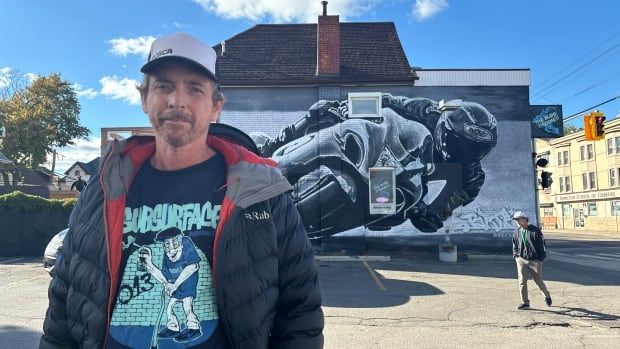 A man stands in front of a mural that depicts a biker.