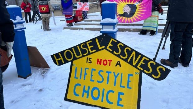 People stand outside a building. There is snow on the ground. Homemade signs and banners read "Houselessness is not a lifestyle choice" and "housing is a human right."