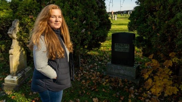 A woman with long red hair and a black vest stands beside a gravestone in the fall