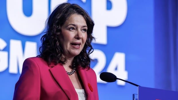 A woman, Danielle Smith, stands at a podium. There is a large blue background behind her that says "UCP AGM 2024" in large, white letters. She has dark hair and is wearing a bright pink pantsuit. 