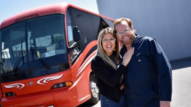 A red bus and a man and a woman stand in front of it smiling.