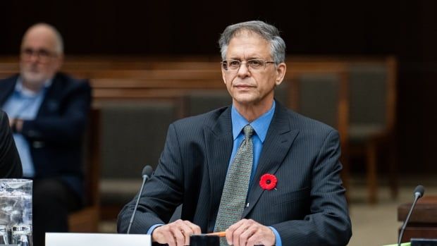 A man in a suit sits at a desk in front of a microphone.