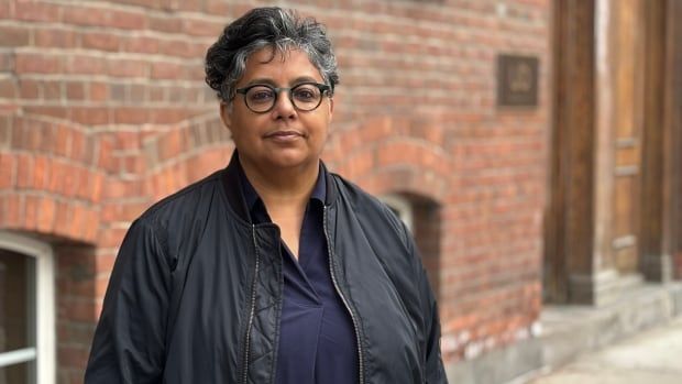 A woman with glasses and black coat looks at the camera in front of a brick wall.