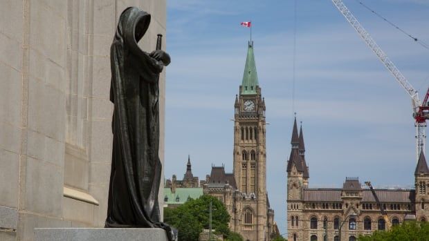 A statue of a cloaked figure, holding a sword, stands at the front of a building. The spires of other buildings are visible in the background. 