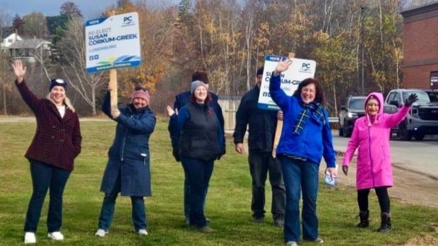 Seven people bundled up in winter clothes and holding campaign signs outside a Tim Horton's drive thru. 