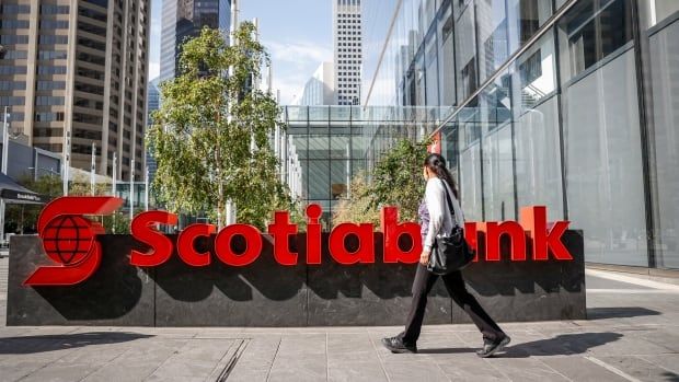 A woman walks past the red letters saying "Scotiabank" on a little raised platform on a city street. 