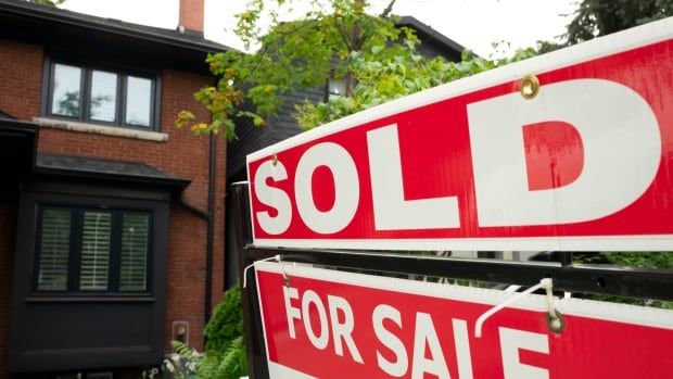 A view of a red sign that says "sold" with a brick house and trees in the background.