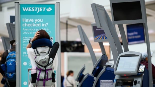 A woman holds her head in her hands while looking at a WestJet sign at the Toronto airport.