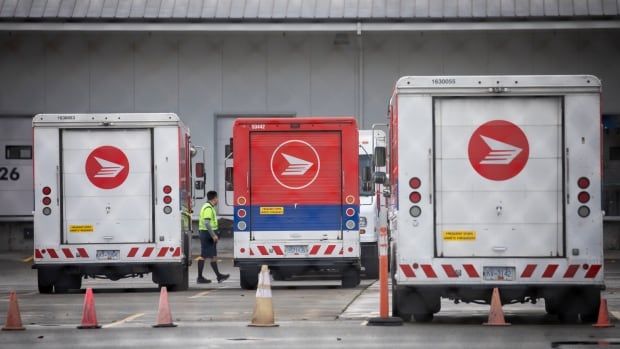 A worker in green high-viz clothing is seen walking between red-and-white delivery trucks at a mail sorting facility.