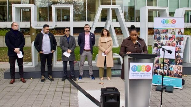 A line of people stand outdoors in front of a sign spelling out "Hamilton." One is at a podium speaking into a mic. 