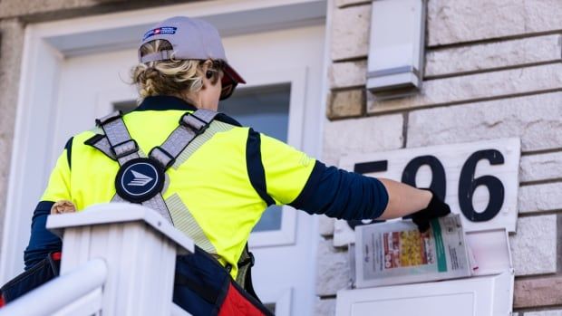 The back of a mail carrier who is wearing a baseball cap is shown. They are extending their right arm toward a mail box.