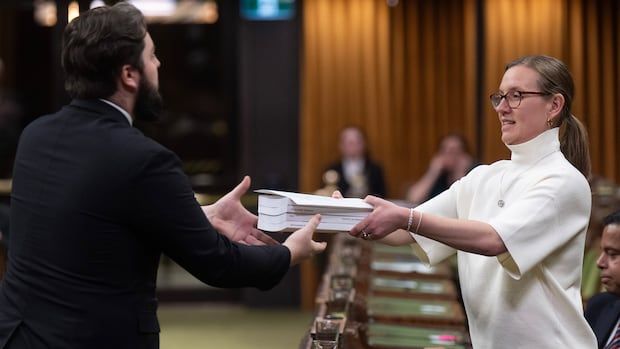 A woman hands a man a stack of books in the House of Commons.