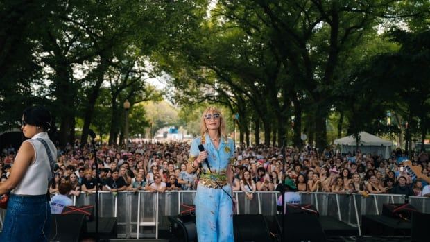 A musician, who has her back turned to the audience, looks at the camera. Behind her are thousands of spectators.