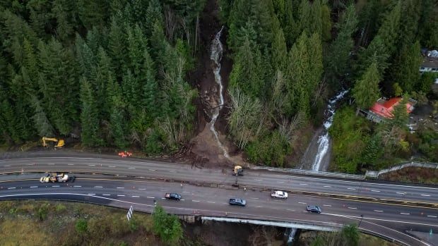 An aerial view of a highway that has had mud and debris over on one side of it, with a landslide clearly visible along a slope.