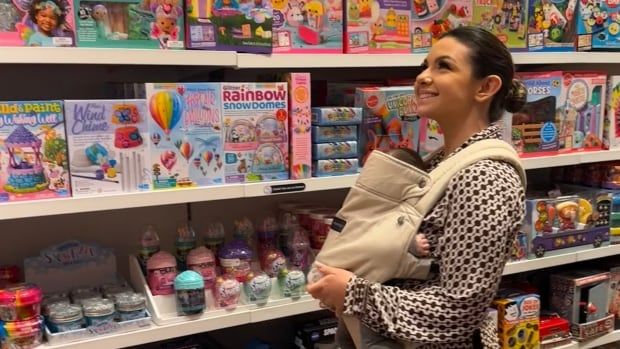 A woman stands in the toy aisle of a store with a baby strapped to her chest. 