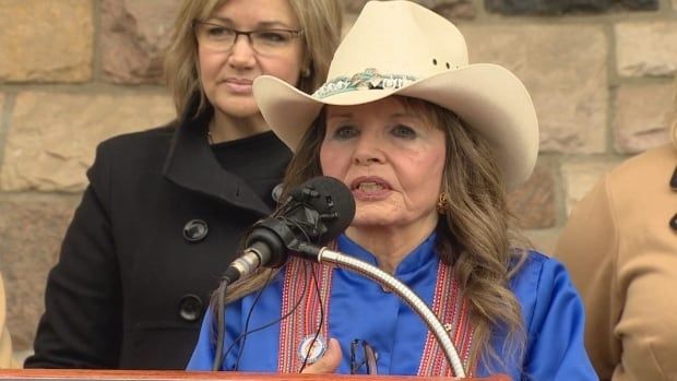 A woman wearing a vibrant blue shirt speaks at a microphone. She is wearing a Métis sash around her shoulders. She is wearing a light-coloured hat with a brim. 