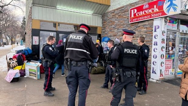 A group of police officers in dark uniforms stands outside a building with a pile of belongings in the background