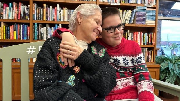 A woman and a boy hold on to each other. The woman's eyes are closed and her head is leaning on the boy's shoulder. The boy is looking directly at the camera, smiling. 